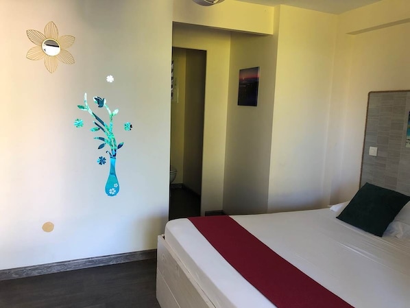 Habitación de hotel con cama, una manta roja, espejo montado en la pared y un adhesivo mural con un jarrón y flores.