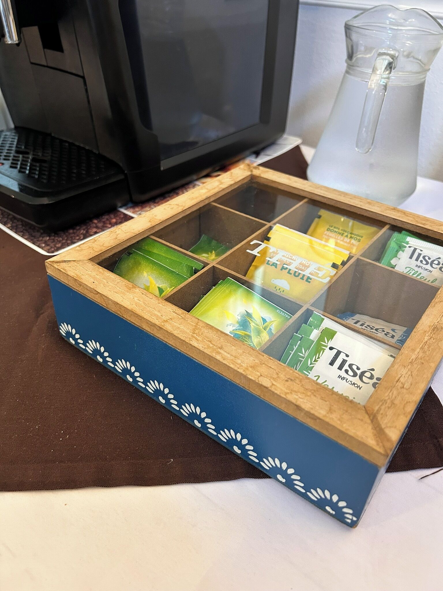 A small wooden box with compartments containing various items, placed on a table next to a computer and a pitcher.