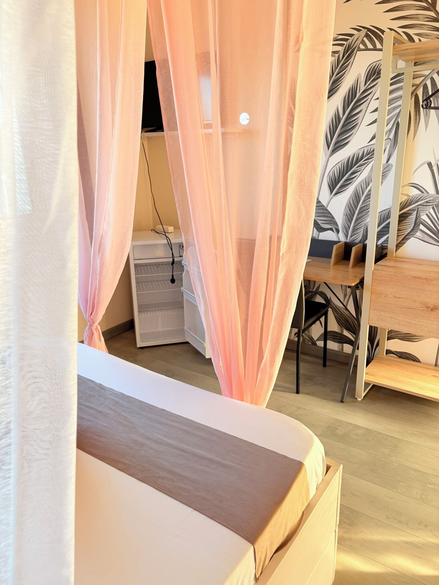 A bedroom with a bed, a desk, and a chair. There is a wardrobe and a bedside table.