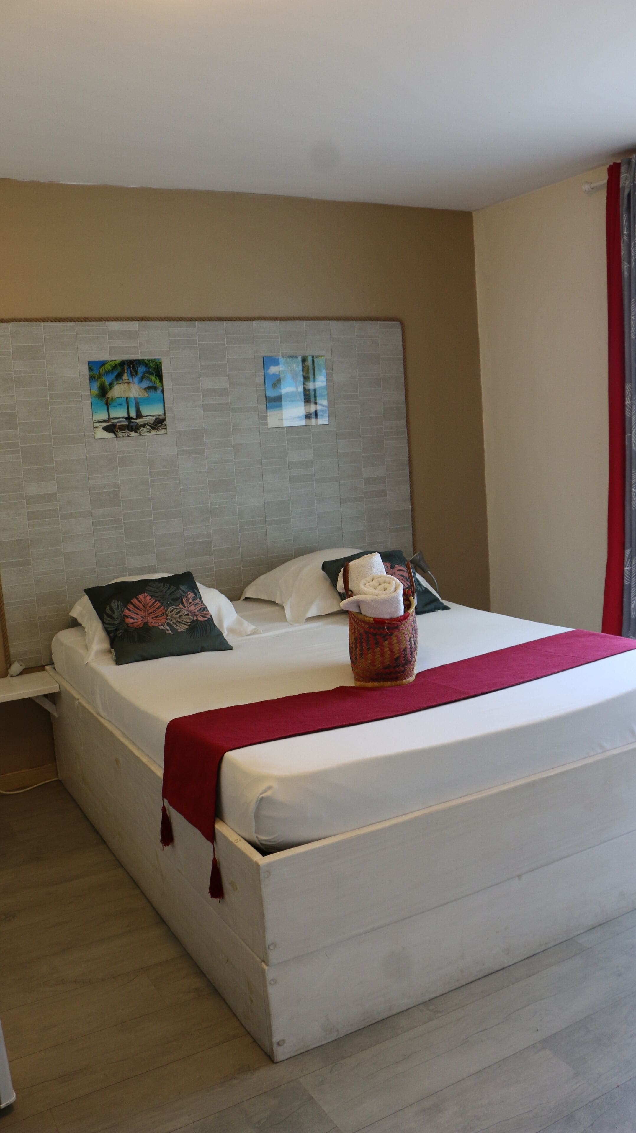 Habitación de hotel con una cama grande, cabecera con obra de arte, manta roja y una cesta tejida con una toalla.