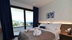 Una habitación de hotel con una cama grande, una mesita de noche con lámpara y vistas a un paisaje urbano a través de un ventanal amplio.