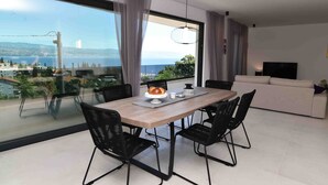 Un comedor moderno con un ventanal grande, una mesa de madera, sillas negras y vistas al mar y al paisaje urbano.