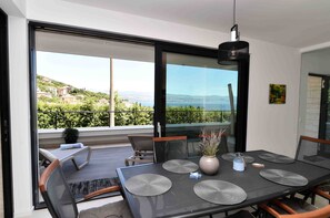 Un espace de repas moderne avec une table dressée pour quatre personnes, de grandes fenêtres offrant une vue sur une colline et la mer, et un luminaire suspendu au-dessus.