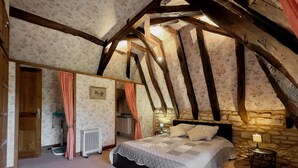 A bedroom with a sloped ceiling, wooden beams, a bed with a quilt, a radiator, a small table with a lamp, and a stone wall. - Gites de Charme 18 Personnes Avec Tennis Dans un Parc de 2 ha . Idéal Familles (Montignac-Lascaux)
