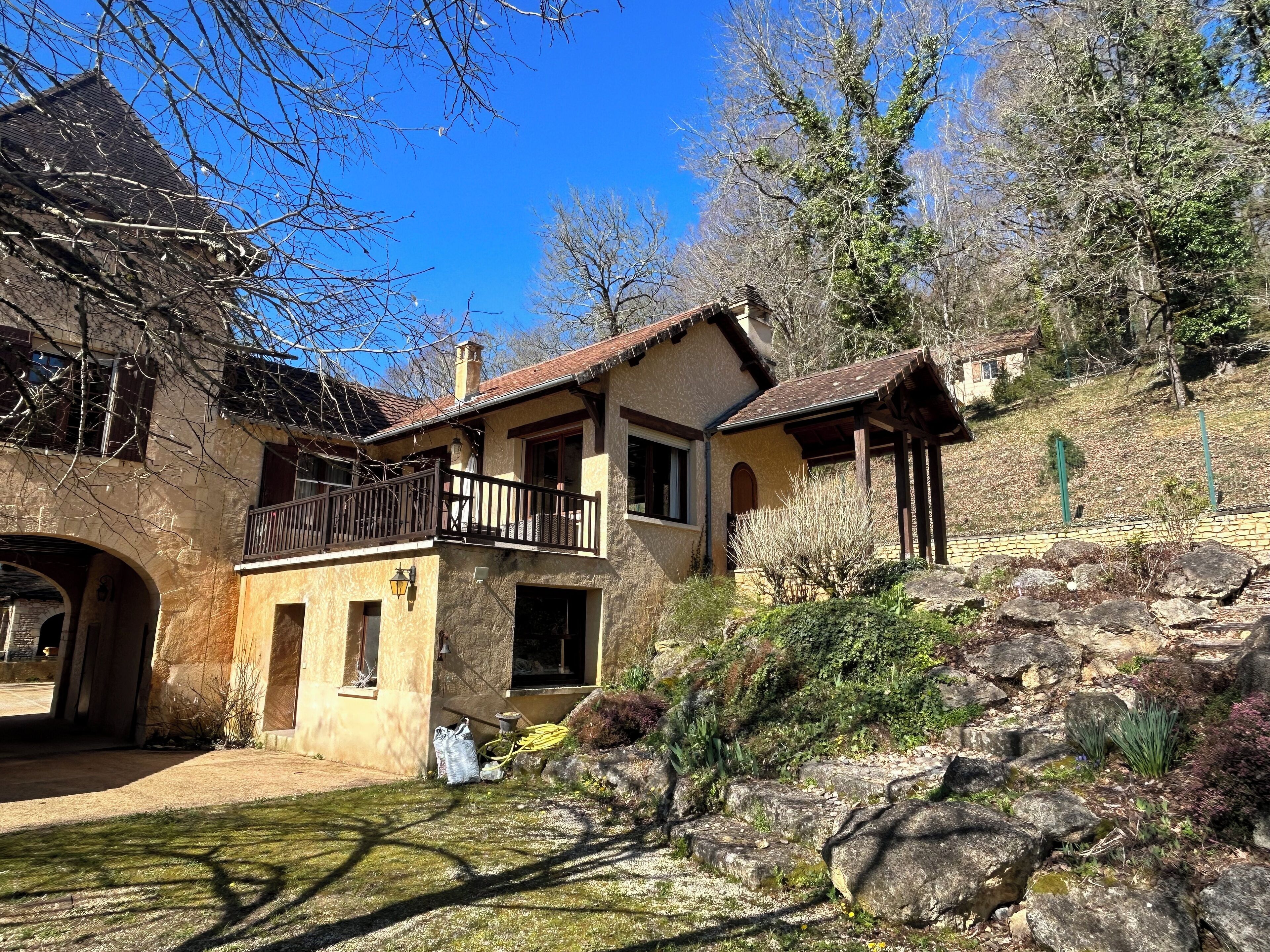 Une maison en pierre à deux étages, dotée d’un balcon, entourée d’un jardin rocheux et d’arbres.