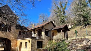 A two-story stone house with a balcony, surrounded by a rocky garden and trees. - Gites de Charme 18 Personnes Avec Tennis Dans un Parc de 2 ha . Idéal Familles (Montignac-Lascaux)