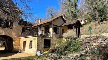 Une maison en pierre à deux étages, dotée d’un balcon, entourée d’un jardin rocheux et d’arbres.