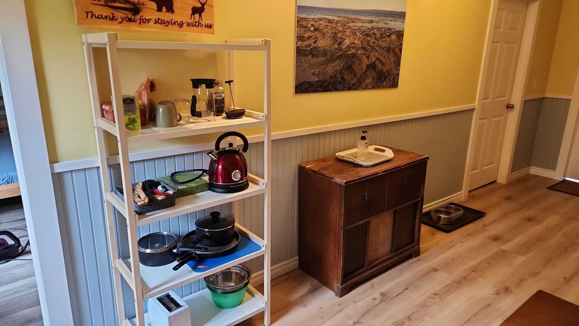 Une cuisine avec une étagère blanche où sont rangés des ustensiles de cuisine, une bouilloire rouge et un tableau représentant un paysage rocheux accroché au mur.