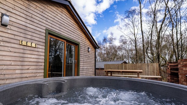 A wooden cabin with a hot tub in front, a wooden bench, and a wooden fence in the background.