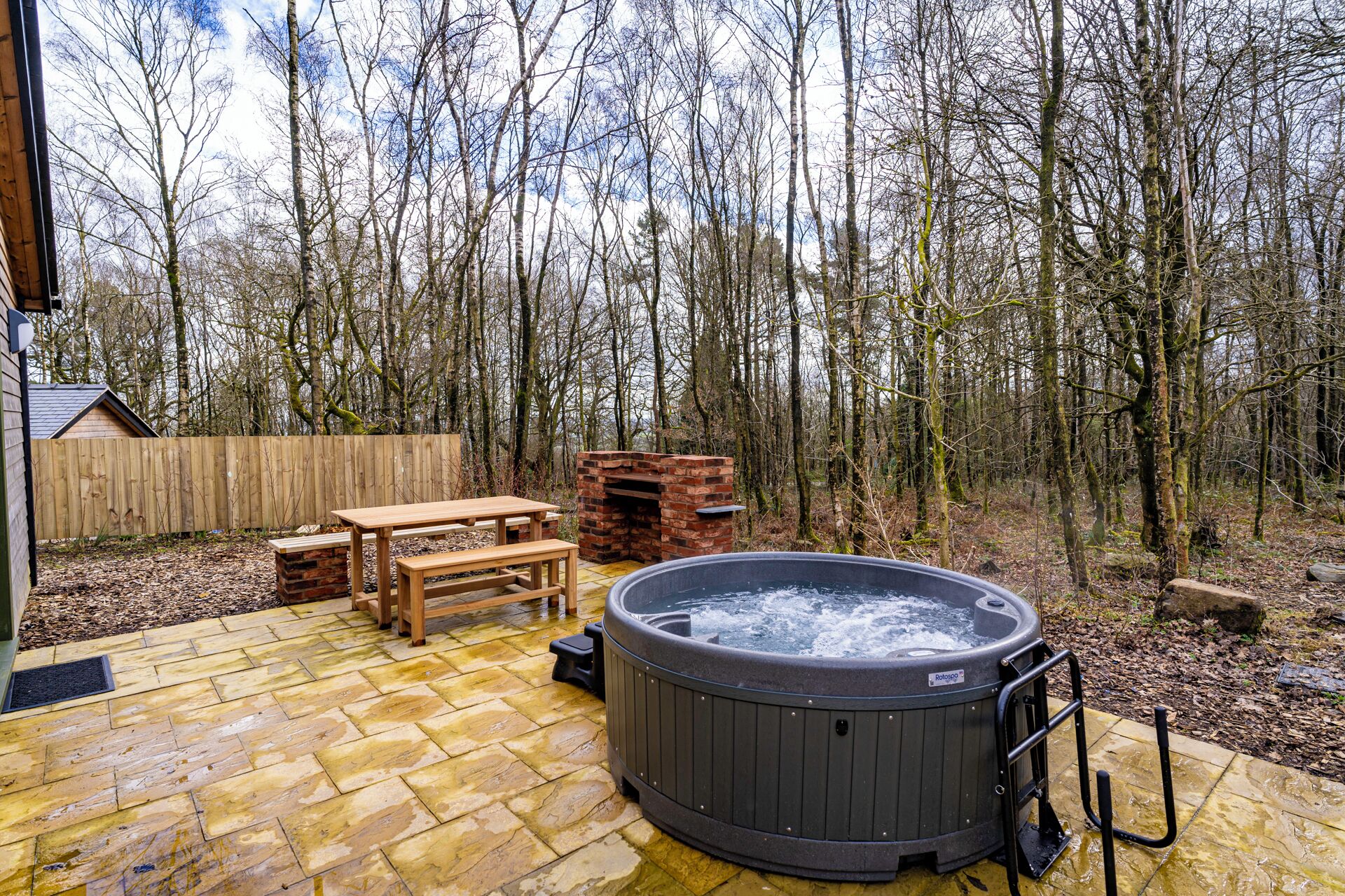 A hot tub in a forested area with a wooden deck, a brick oven, and a picnic table.