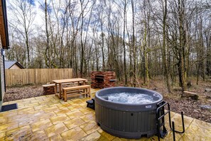 Ein Whirlpool in bewaldeter Landschaft mit einer Holzterrasse, einem Backofen aus Ziegeln und einem Picknicktisch.