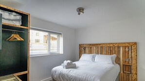 A bedroom with a wooden headboard, a bed with white linens, a window with blinds, and a wooden wardrobe with hangers.