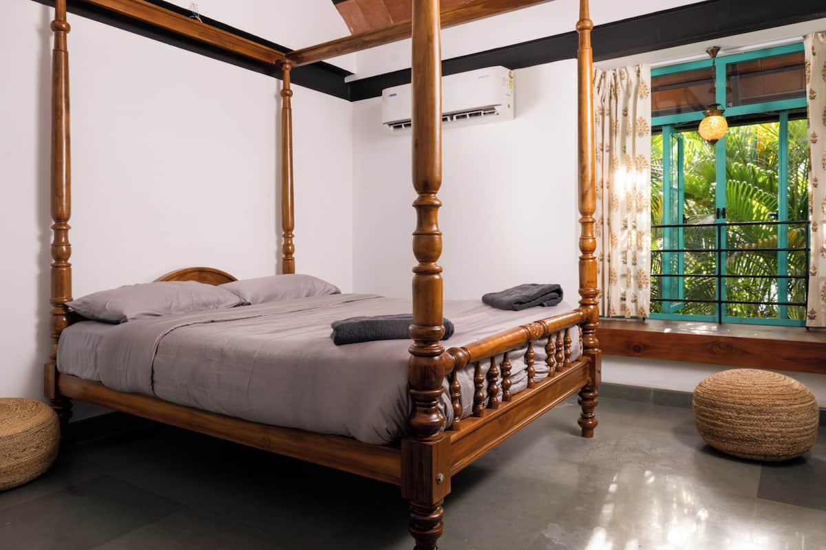 A four-poster bed with a wooden frame and a grey bedspread, a window with green shutters, and a ceiling fan.