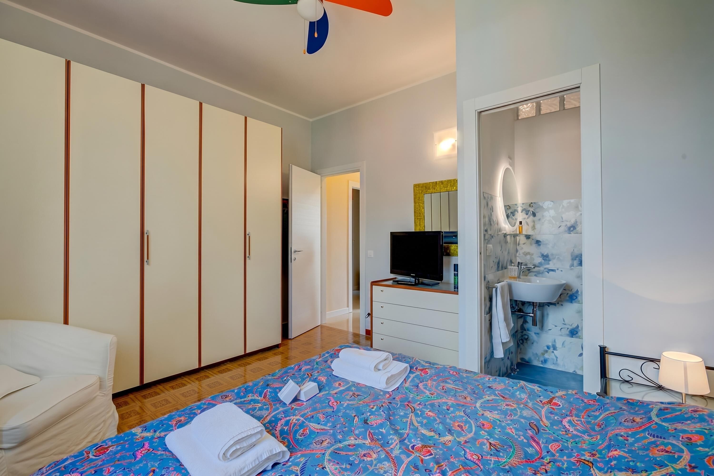 A bedroom with a bed, a TV, a wardrobe, a bathroom with a sink and mirror, and a door leading to another room.
