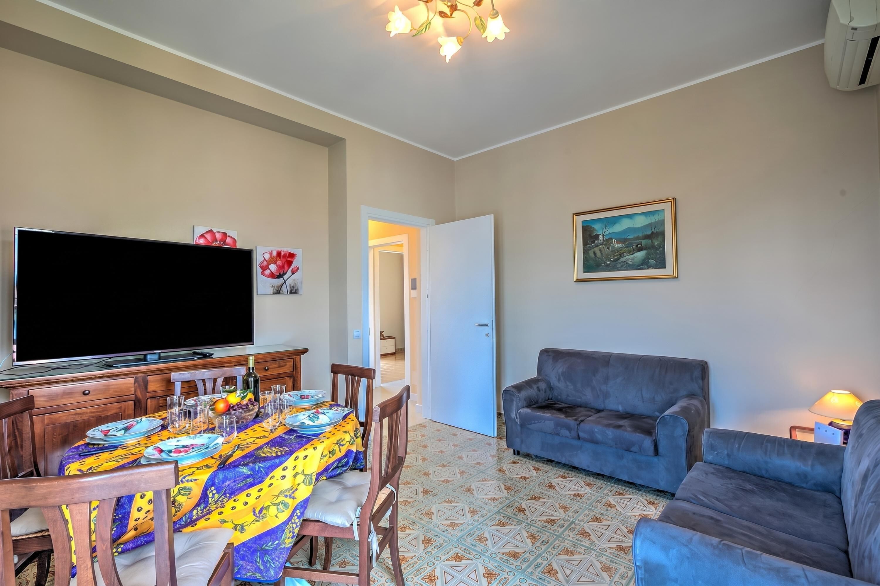 Un comedor con una mesa dispuesta para cuatro personas, un televisor, un sofá y un cuadro en la pared.