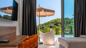 A modern bedroom with a large window, a flat-screen TV, a wooden table, a unique orange chair, and a bed with a view of a green landscape. - Luxury Villa Morello Korcula 1 with Heated Pool (Korcula)