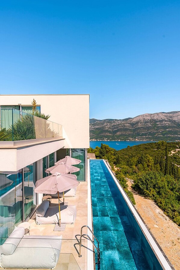 A modern infinity pool with lounge chairs and umbrellas, overlooking a mountainous landscape and a body of water. - Luxury Villa Morello Korcula 2 with Heated Pool (Korcula)