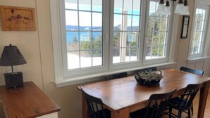 A dining area with a wooden table and four chairs, large windows with a view, and a lamp on a side table. - Beautiful oceanfront cottage (Guysborough)