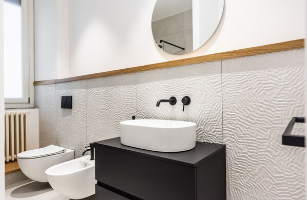 A modern bathroom with a white sink, black faucet, and a round mirror above it. There is a black vanity and two white bidets. - Agata Apartment with Balcony by Wonderful Italy (Catania)
