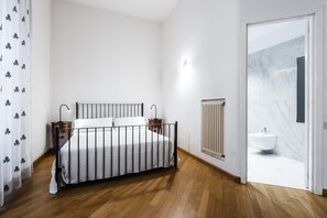 A bedroom with a wooden floor, a bed with a metal frame, a bedside table, a radiator, and a bathroom with a marble sink. - Agata Apartment with Balcony by Wonderful Italy (Catania)