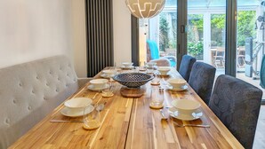 Un coin repas avec une table en bois dressée pour quatre personnes, un luminaire suspendu moderne et une vue sur l’extérieur grâce à de grandes fenêtres.