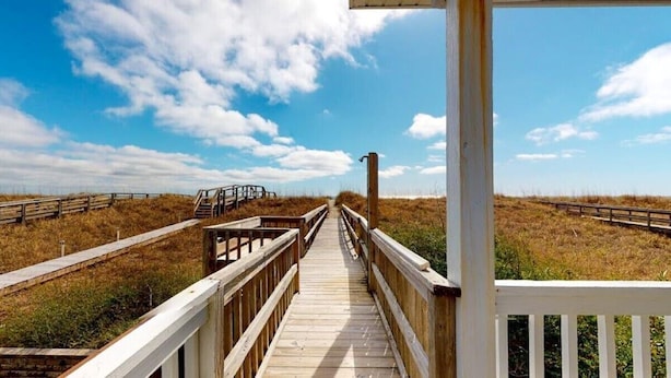 Une promenade en bois mène à une plage, avec une rambarde en bois d’un côté et une clôture blanche de l’autre.