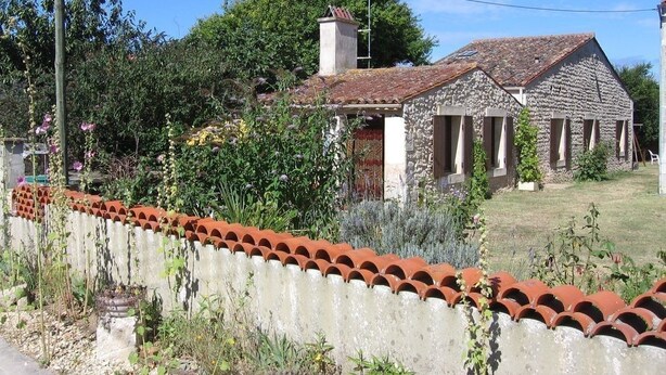 Una casa de piedra con techo de tejas, rodeada por un jardín con diversas plantas y una valla blanca con tejas rojas.