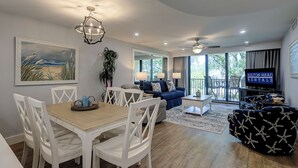 Una sala de estar moderna con comedor, un sofá azul, televisor y balcón con mobiliario de exterior.