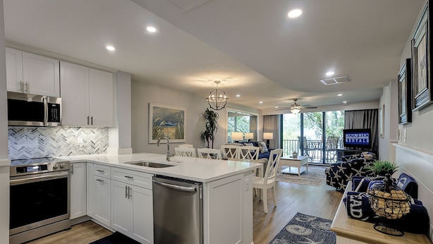 Una cocina moderna con gabinetes blancos, electrodomésticos de acero inoxidable y una isla central. Cuenta con un comedor con una araña y un televisor que muestra 'Hilton Head Rentals'.