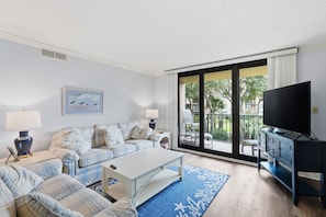 A living room with a sofa, coffee table, and a television. There is a large window with a view of a balcony and greenery outside. - 4203 Island Club - Folly Field - 2 Bed 2 Bath Sleeps 5 - Hilton Head Rentals (Hilton Head Island)