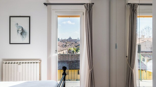 Een slaapkamer met een bed, een radiator, een lijstwerk en uitzicht op een stadsgezicht door een raam met gordijnen.