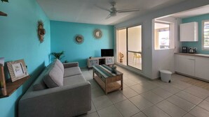 A living room with a sofa, coffee table, and a TV. There is a large window and a ceiling fan. - Le Soleil Bleu, Charmant T2 à Proximité des Plages, Restaurants et Commerces (Le Gosier)