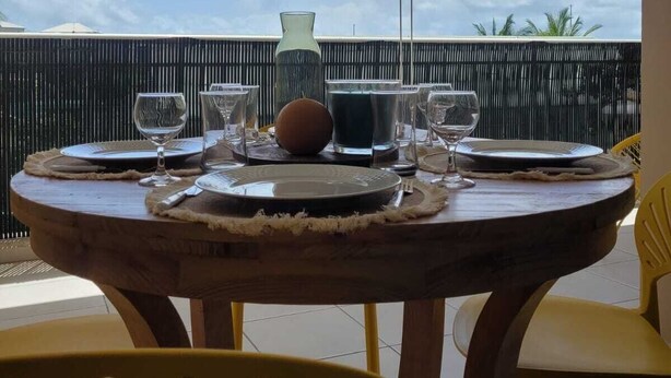 A round wooden table set for dining with plates, glasses, and a pitcher, positioned on a balcony with a view of the sky and palm trees. - Le Soleil Bleu, Charmant T2 à Proximité des Plages, Restaurants et Commerces (Le Gosier)