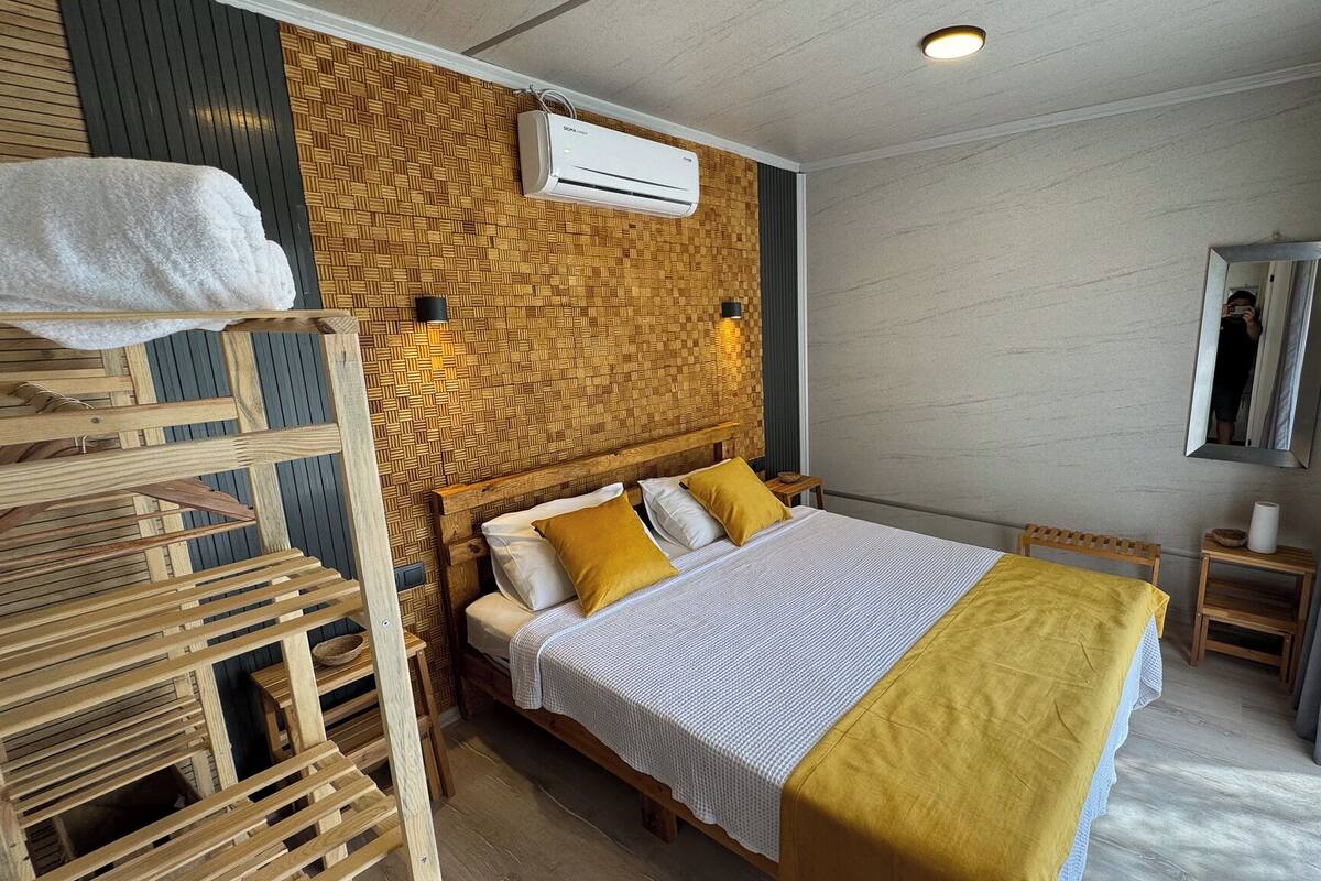 A modern hotel room with a wooden headboard, a bed with white and yellow bedding, a wooden ladder, and a wall-mounted air conditioner.