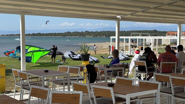 A covered outdoor seating area with tables and chairs, overlooking a beach with people and kites.