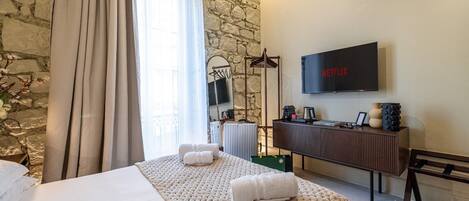 A hotel room with a stone wall, a bed with a beige quilt, a bedside table with a lamp, a TV on the wall, and a window with curtains.