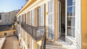 A building with a balcony, multiple windows with shutters, and a visible interior with furniture. - Naomi Nice  (Nice)