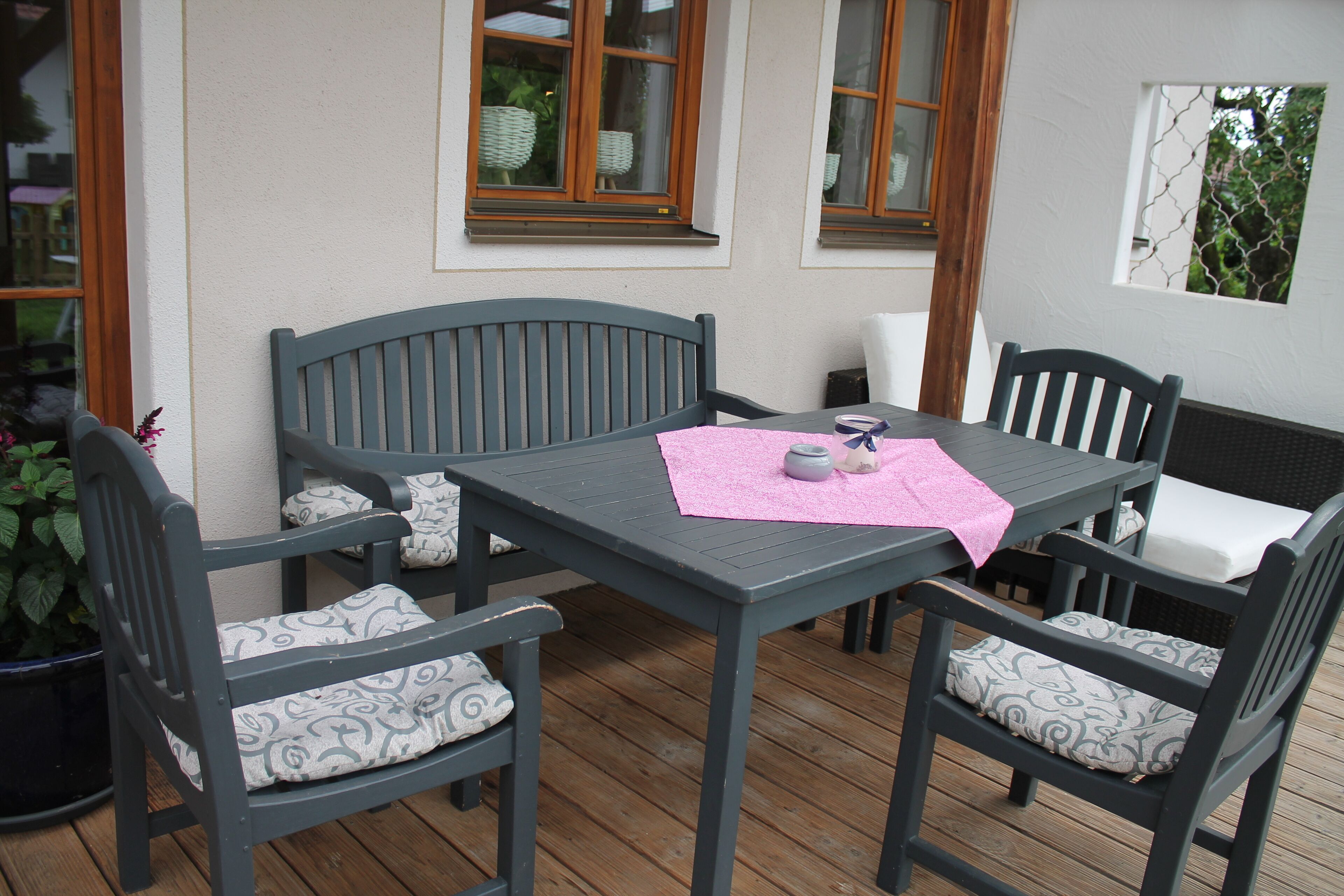 A wooden deck with a table and chairs, a potted plant, and a window with a view of greenery.