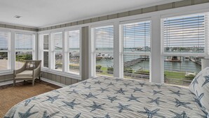 A bedroom with a large bed, a chair, and multiple windows overlooking a marina. - Gorgeous sunset views looking over the waterway! 4BR/2.5Bath (Wrightsville Beach)