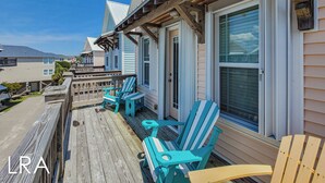 A wooden deck with Adirondack chairs, a covered porch, and a view of neighboring houses. - Melon Camp - Dive into Fun with Pool Access! (Surf City)