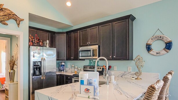 A kitchen with dark wooden cabinets, a stainless steel refrigerator, a microwave, and a marble island with chairs. - Melon Camp - Dive into Fun with Pool Access! (Surf City)