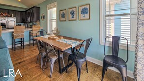 A dining area with a wooden table, metal chairs, and a kitchen in the background. - Melon Camp - Dive into Fun with Pool Access! (Surf City)