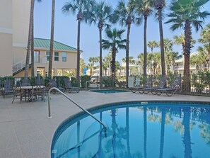 Zona de piscina con sillones y mesas, rodeada de palmeras y un edificio con techo verde.
