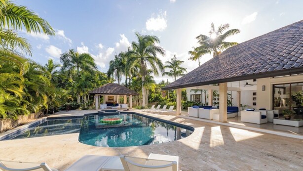 A pool area with lounge chairs, a covered seating area, and a building with a tiled roof.