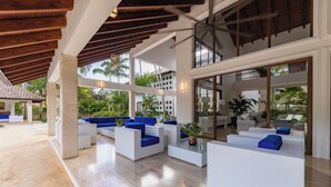 A spacious outdoor lounge area with white sofas and blue cushions, large windows, and a wooden ceiling.