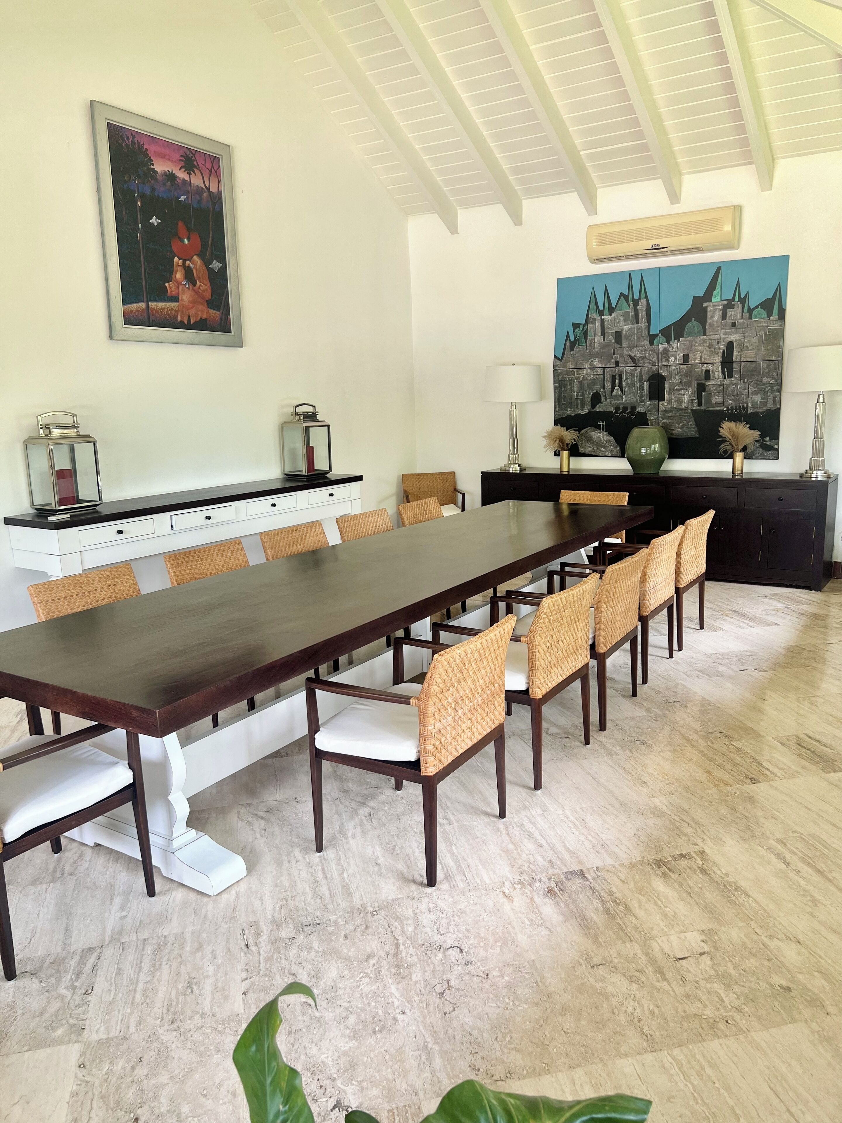 Un comedor con una mesa larga, sillas de mimbre y una consola blanca con objetos decorativos.