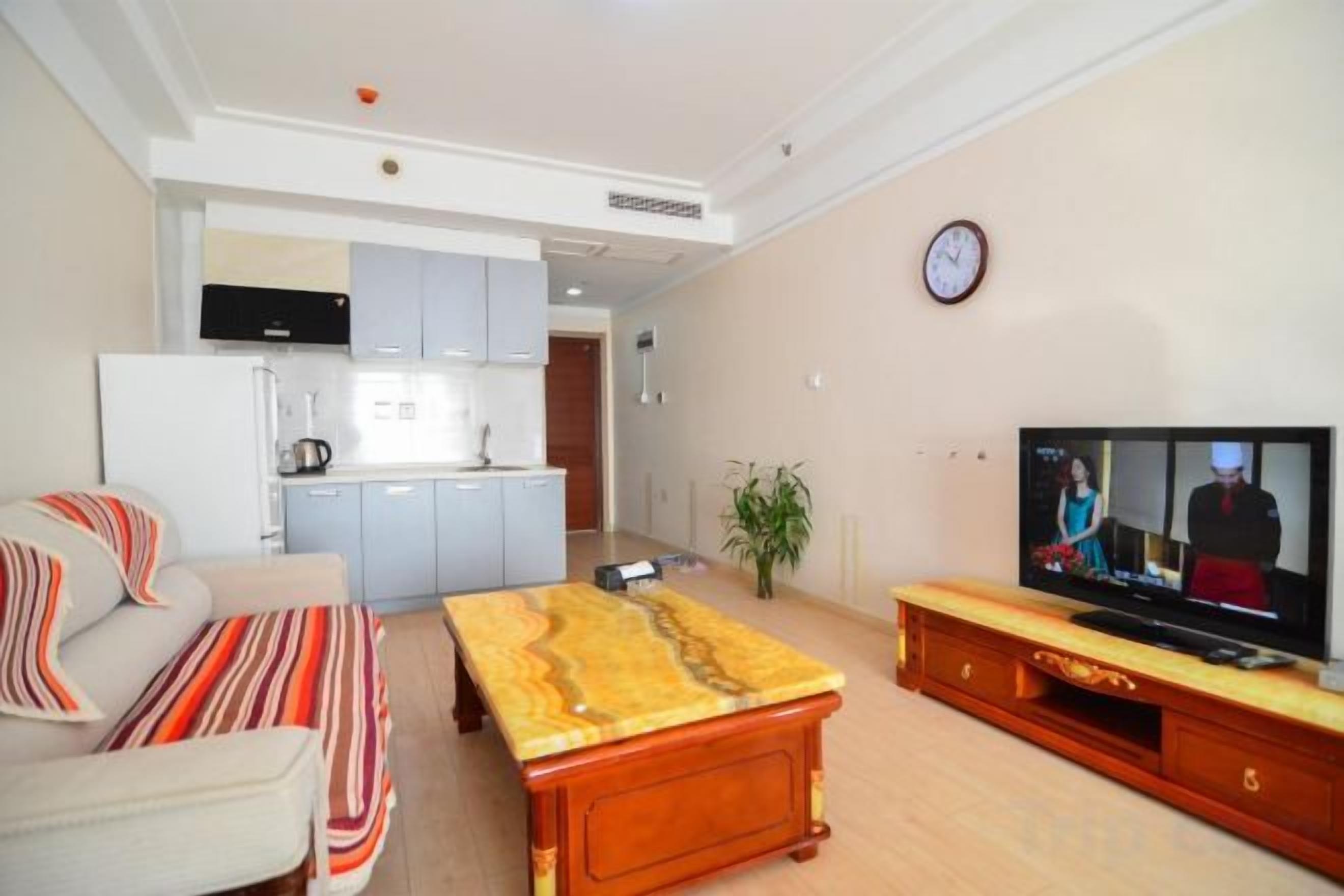 Una sala de estar con sofá, mesa de centro y televisor. Hay una zona de cocina con armarios y refrigerador.