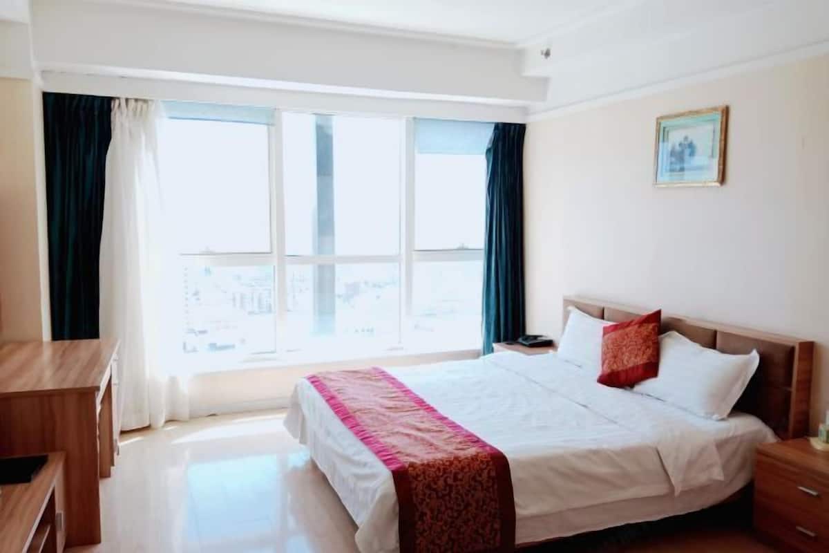Habitación de hotel con un ventanal grande, una cama con ropa de cama blanca y un cojín rojo decorativo, una mesita de noche de madera y un escritorio pequeño.