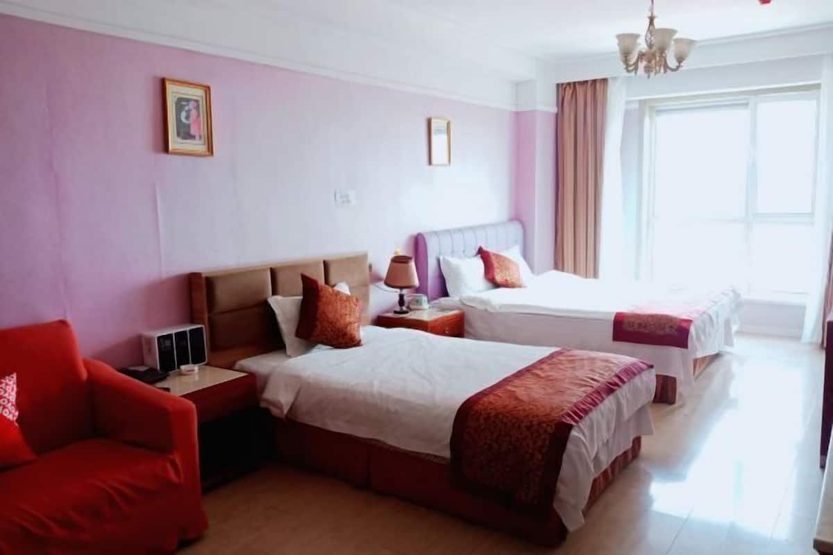 Habitación de hotel con dos camas, un sillón rojo, una mesita de noche con lámpara y un ventanal con cortinas.