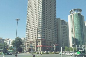 Une vue urbaine avec de hauts immeubles, un ciel dégagé et une rue animée, où l’on voit des véhicules et des piétons.